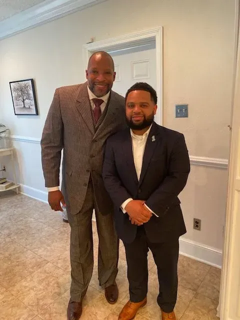 Two men in suits and ties are standing next to each other in a hallway.