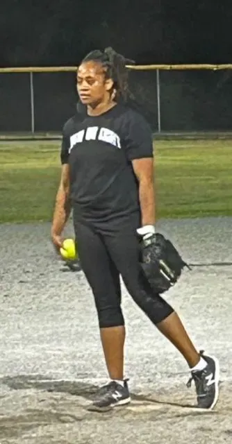 A woman in a black shirt is holding a softball and a glove.