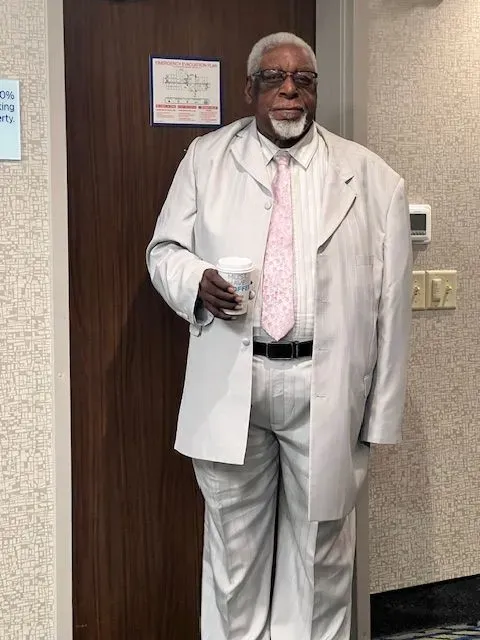 A man in a white suit and pink tie is standing in front of a door holding a cup of coffee.