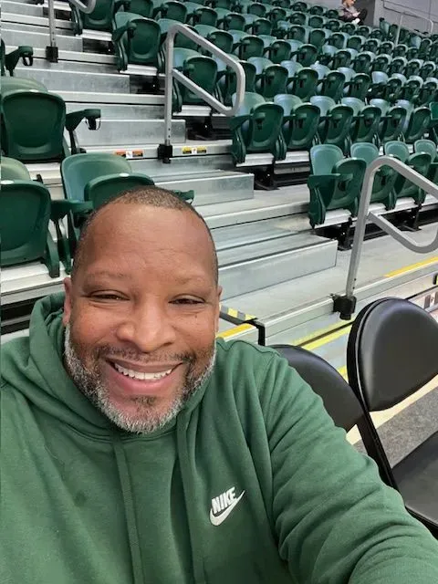 A man in a green nike hoodie is taking a selfie in an empty stadium.