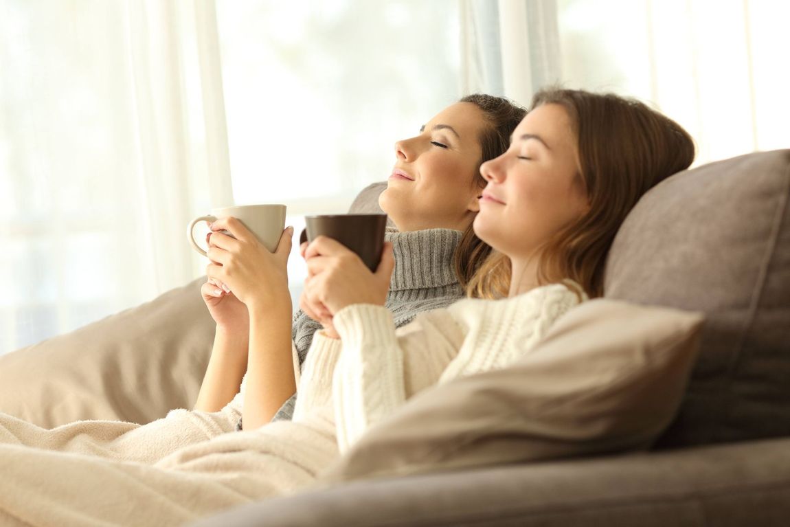 two women are sitting on a couch holding cups of coffee