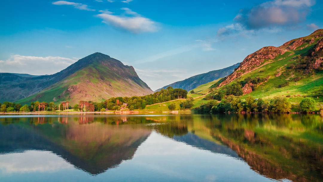 Scenic Lake District landscape with mountains, green grass, and blue skies reflected in water.