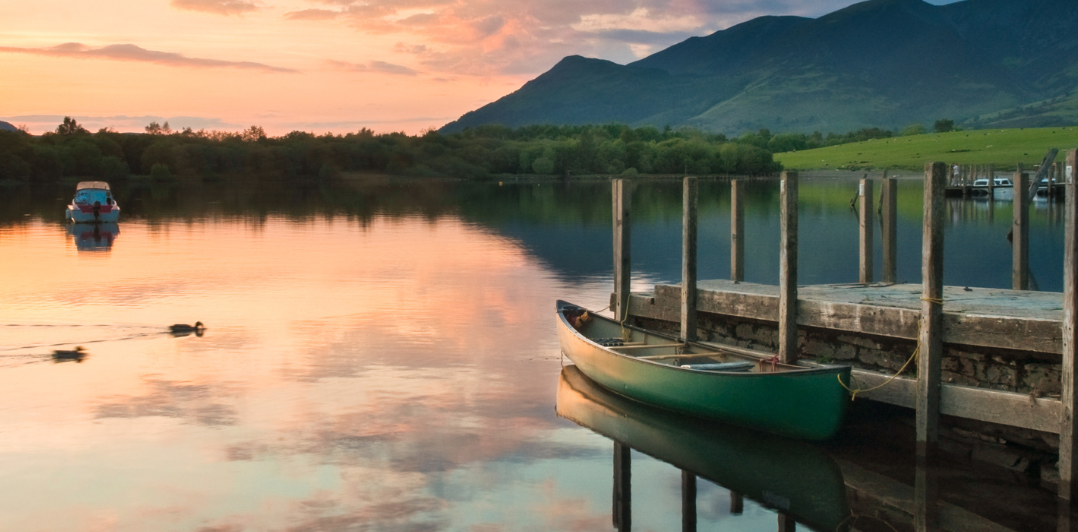Lake Windermere Sunset scene at a lake with a green boat tied to a stone and concrete jetty, mountains and trees silhouette against a pink sky.