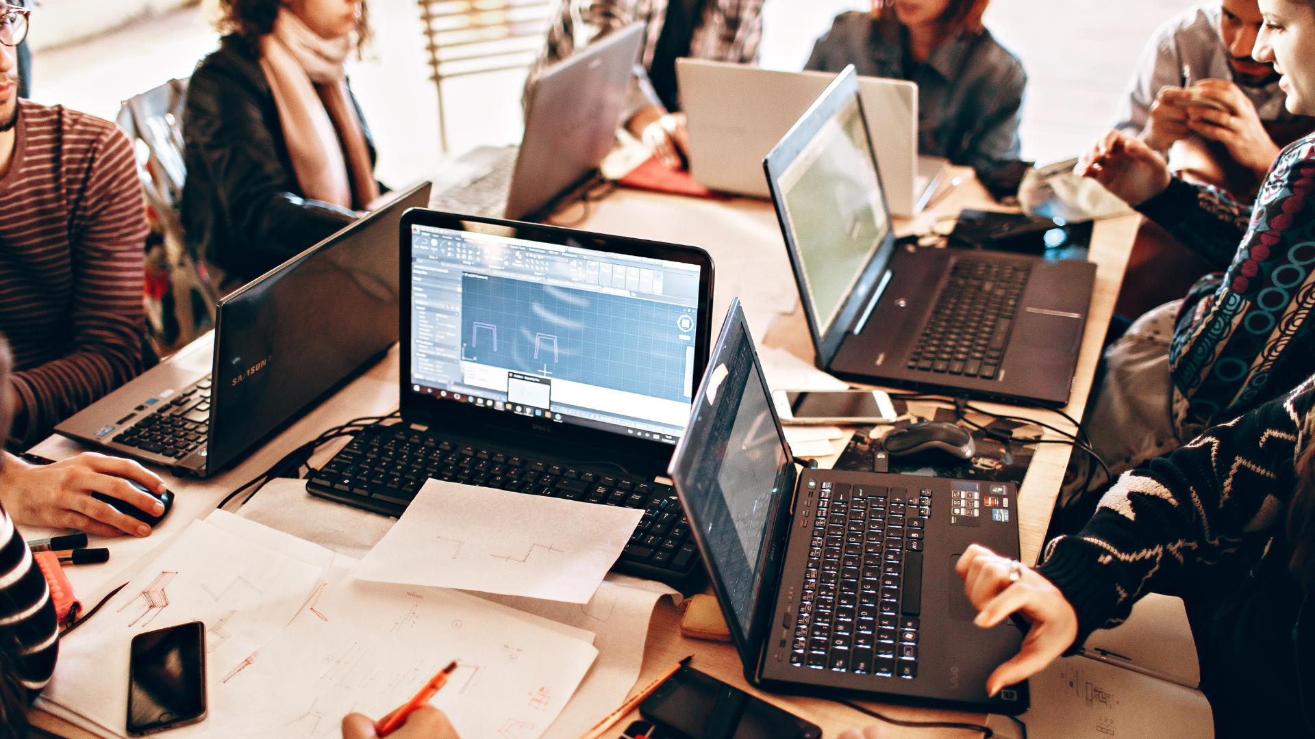 People gathered around laptops at a shared table, collaborating in a bright office workspace.