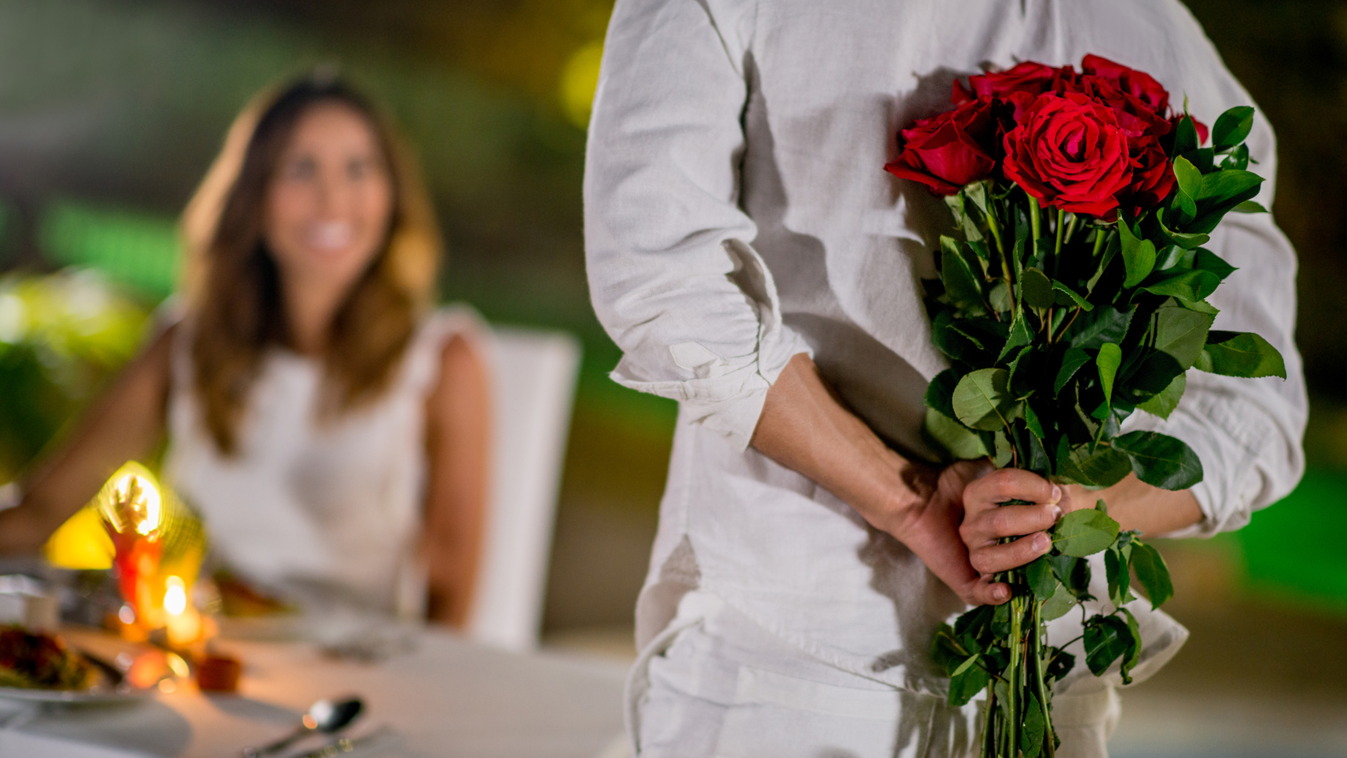 Person holding a bouquet of red roses in a softly lit restaurant, with a smiling diner blurred in the background