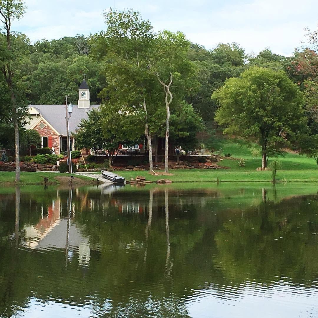 A calm pond reflecting trees and a small house beside a grassy shore