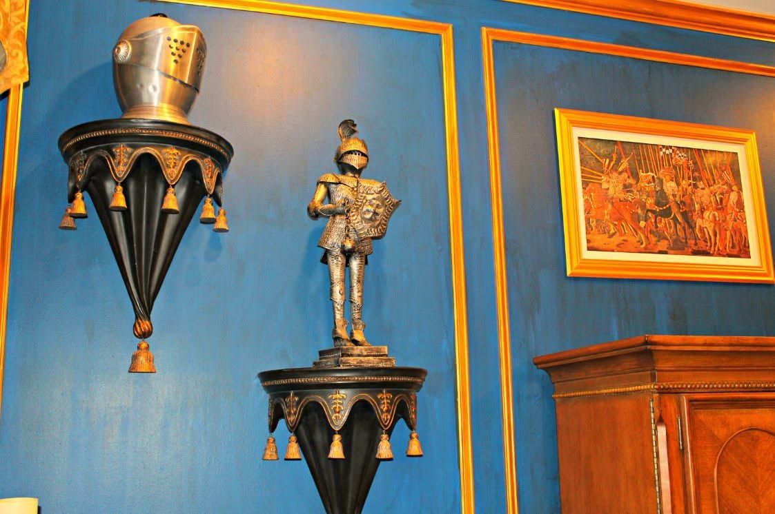 Blue wall with gold trim, two ornate wall sconces, and a framed painting beside a wooden cabinet