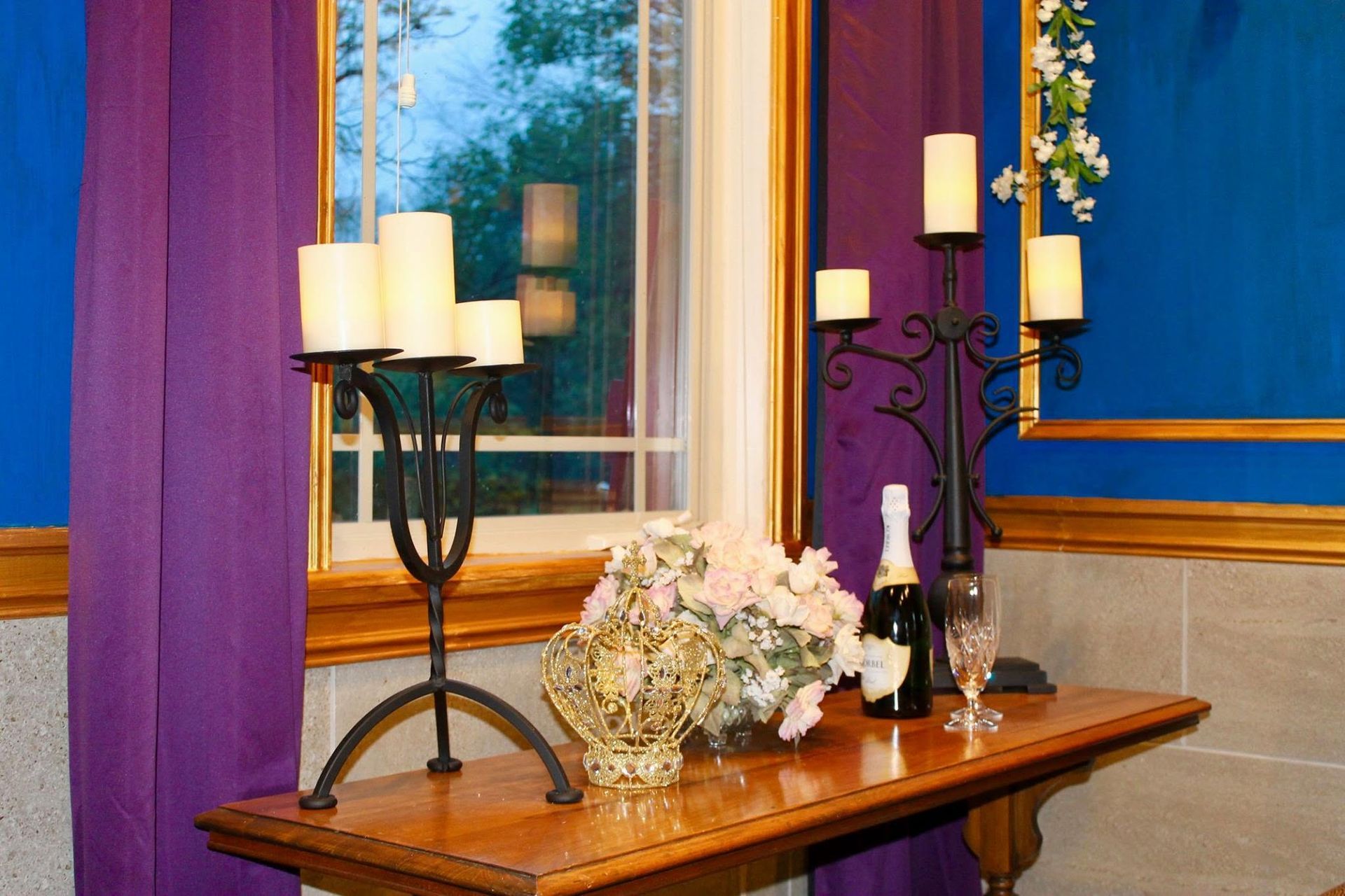 Decorated table by a window with candles, flowers, and a champagne bottle in a purple-draped room