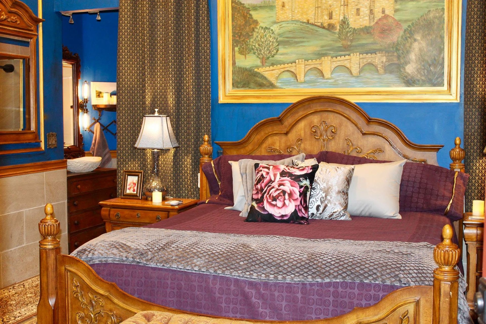Elegant bedroom with carved wooden bed, purple bedding, lamp, and blue walls with artwork
