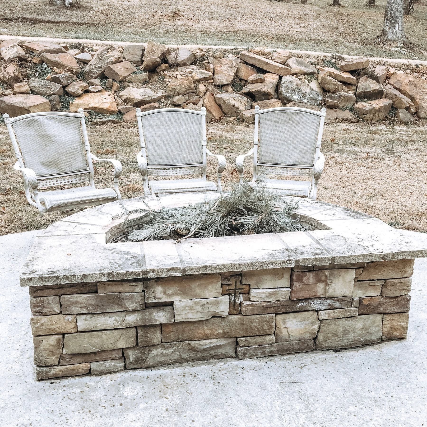 Three white chairs behind a stone fire pit on a gravel patio, with rocks and dry grass in the background