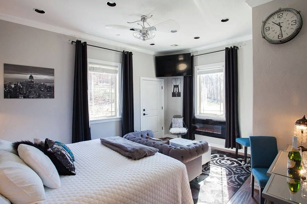Bright bedroom with white bedding, dark curtains, two windows, a blue chair, and a large wall clock