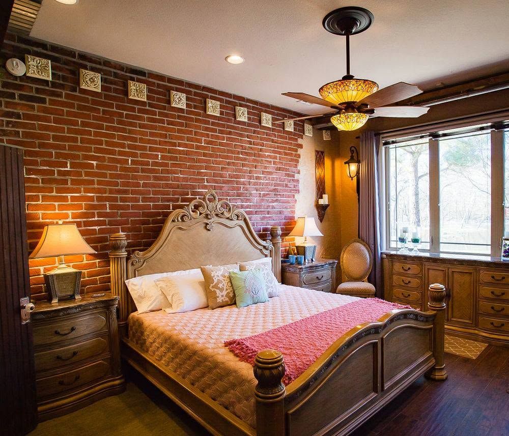 Cozy bedroom with brick accent wall, ornate bed, warm lamps, ceiling fan, and large windows