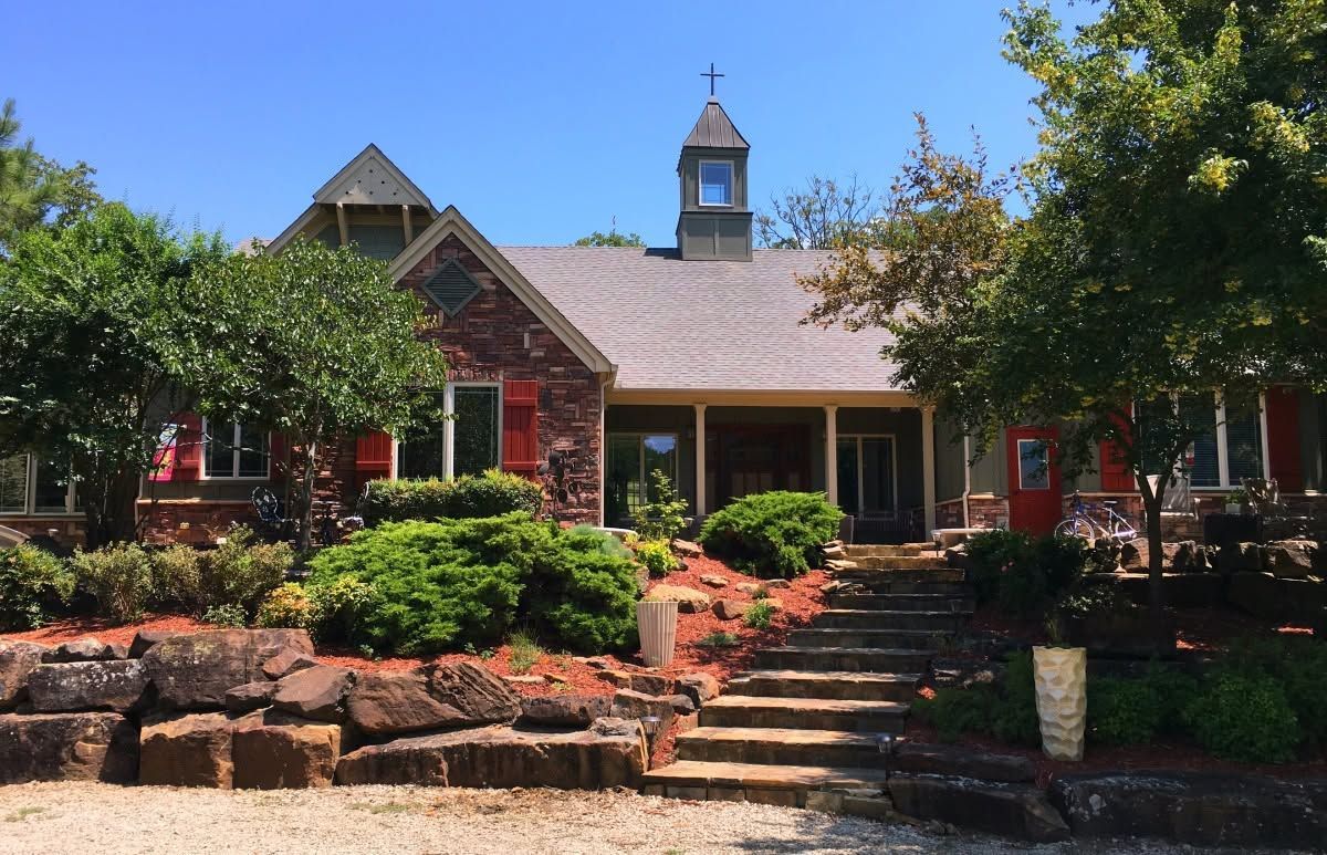 Red-brick house with front porch, stone steps, and landscaped garden under a blue sky