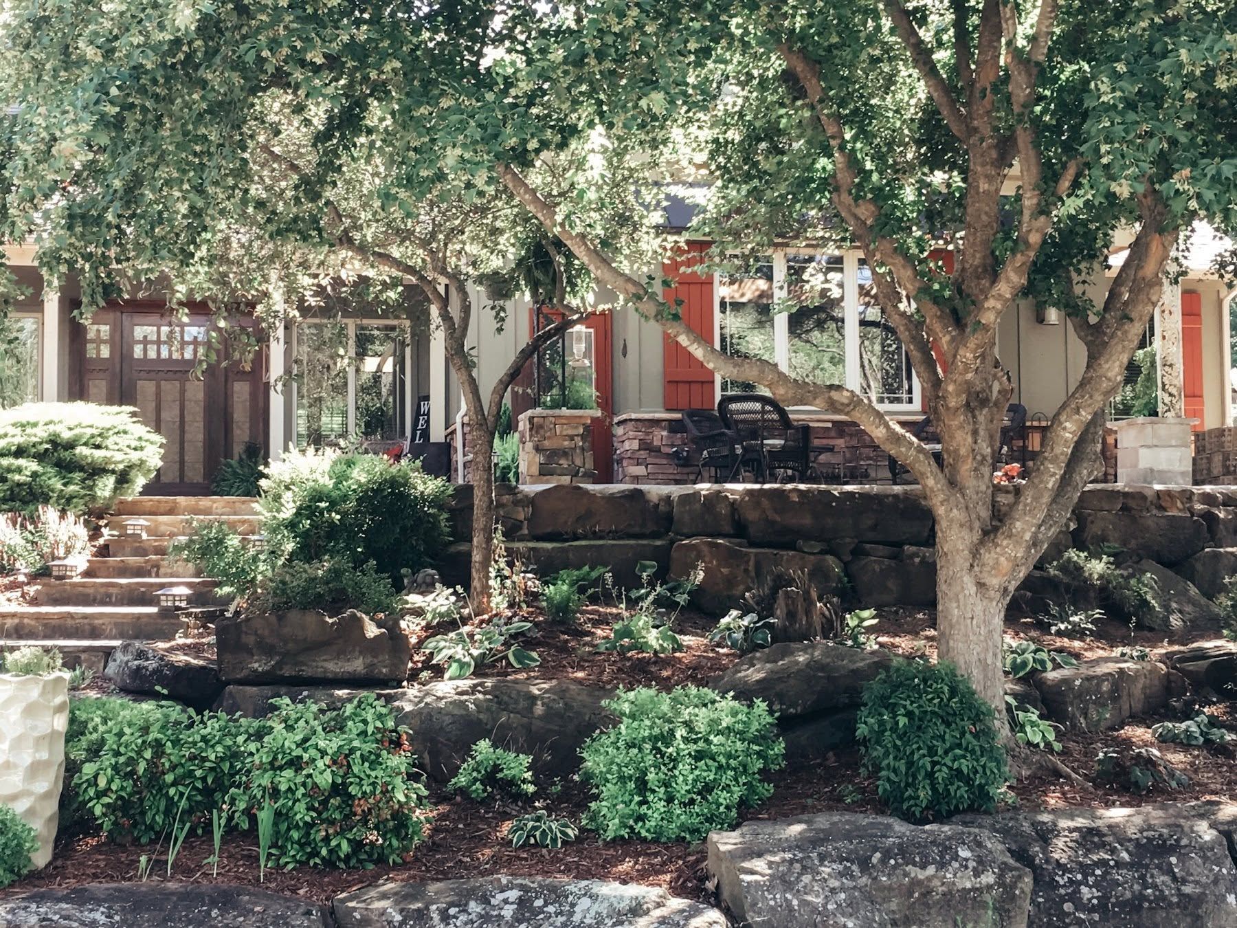 Shaded garden courtyard with stone retaining wall, lush shrubs, and a large tree in front of a house