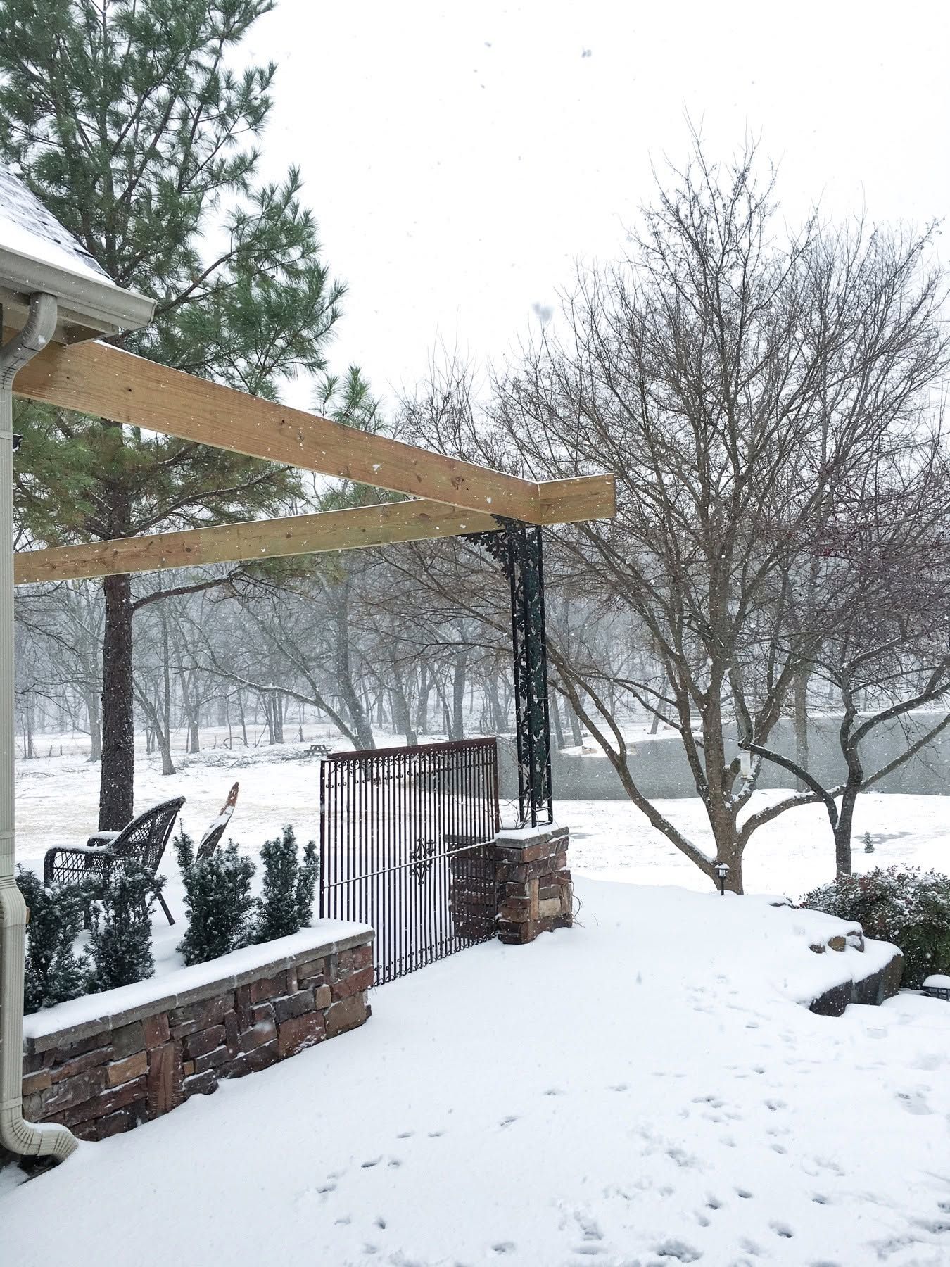 Snow-covered backyard patio with pergola, stone wall, and leafless trees in winter