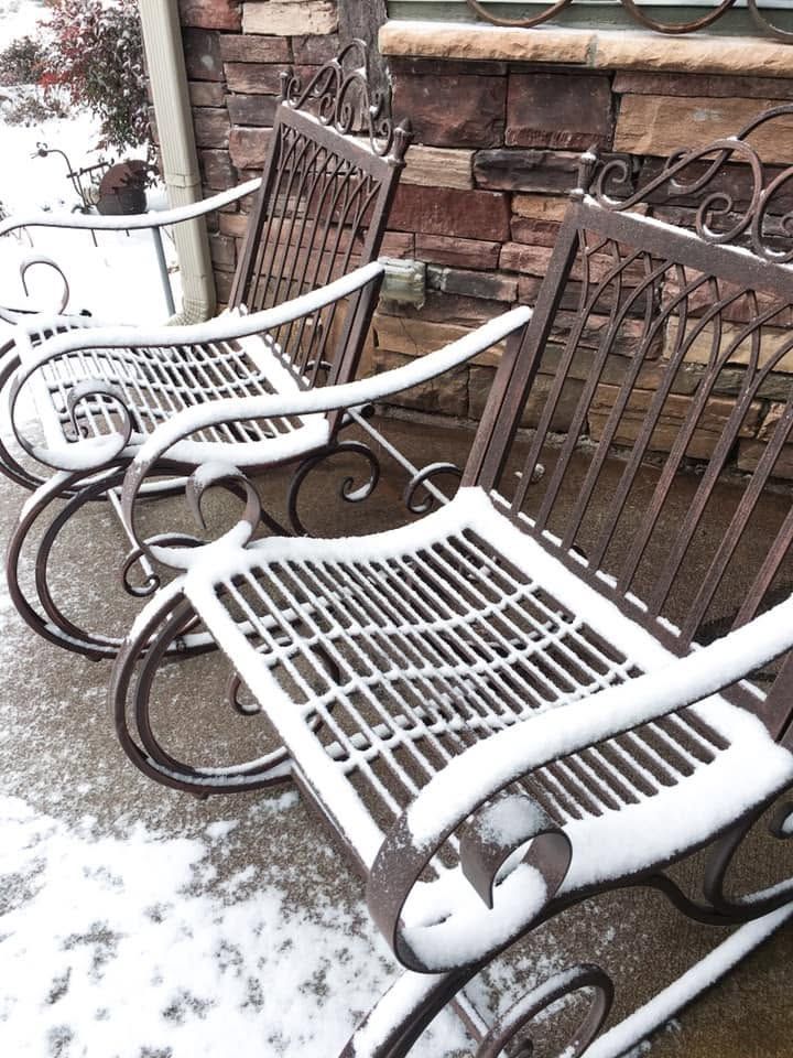 Snow-covered white metal patio chairs beside a brick wall.