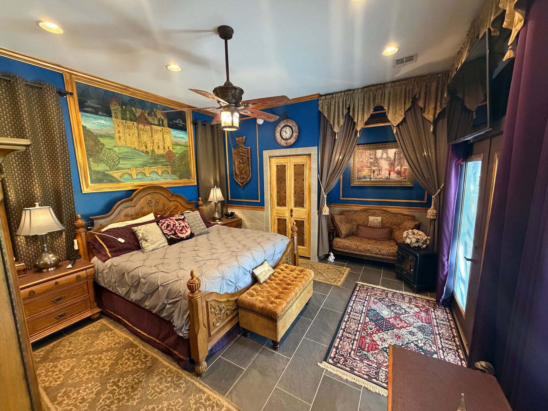 Ornate bedroom with carved wooden bed, purple bedding, blue walls, lamp, and framed artwork