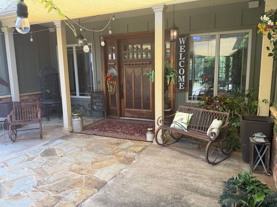 Front porch with wooden door, stone patio, bench, potted plants, and a vertical “WELCOME” sign.