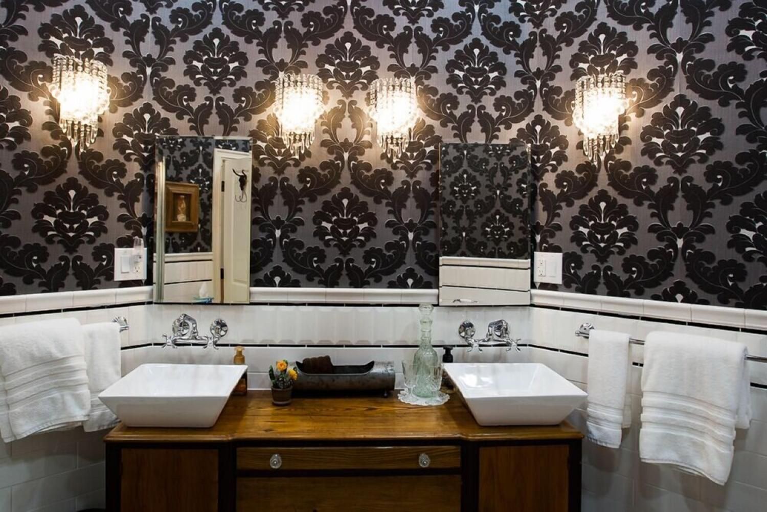 Elegant bathroom vanity with two sinks, large mirror, patterned wallpaper, and wall sconces
