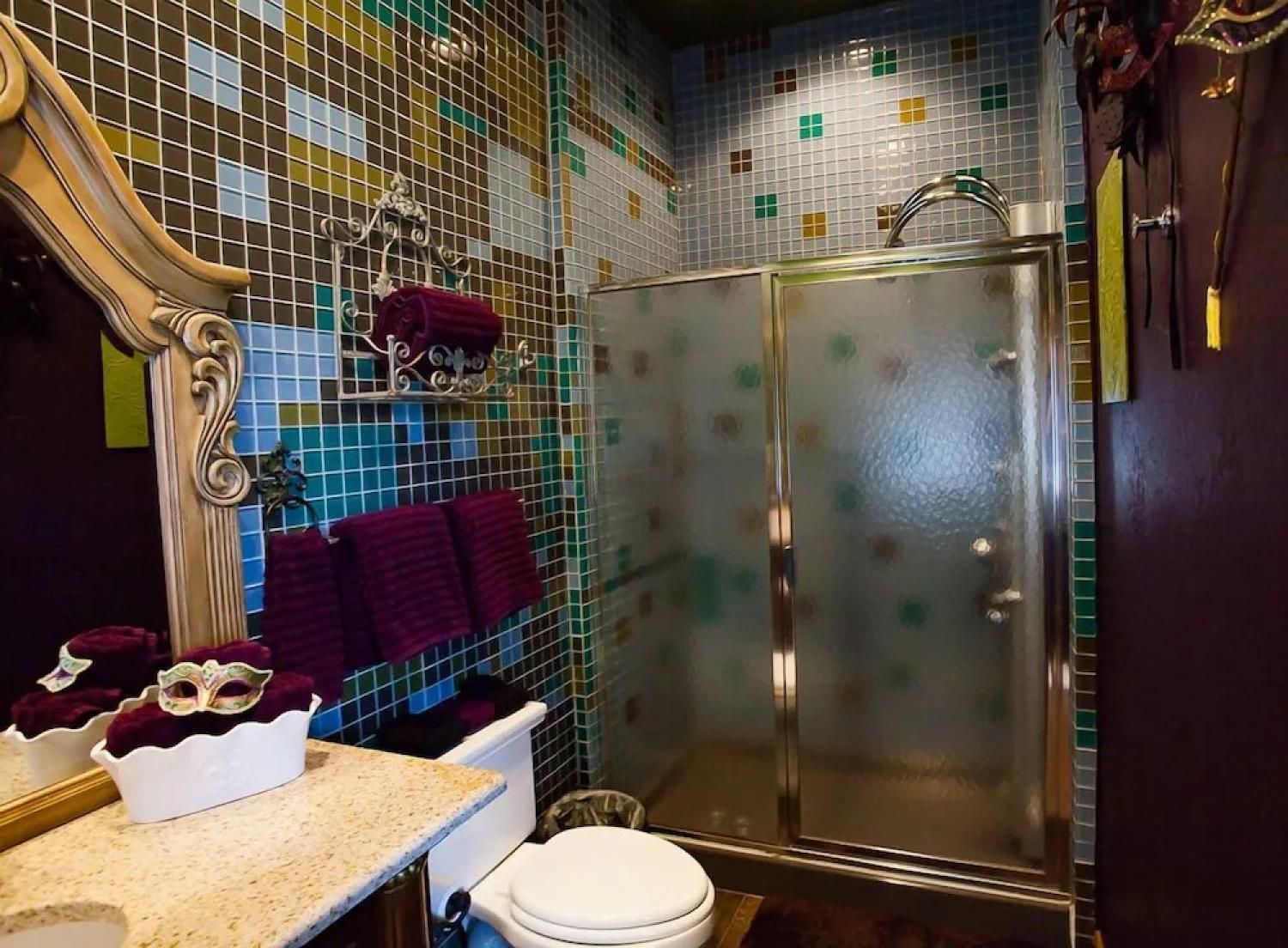 Small bathroom with mosaic tile walls, frosted glass shower, ornate mirror, and toilet beside a sink.