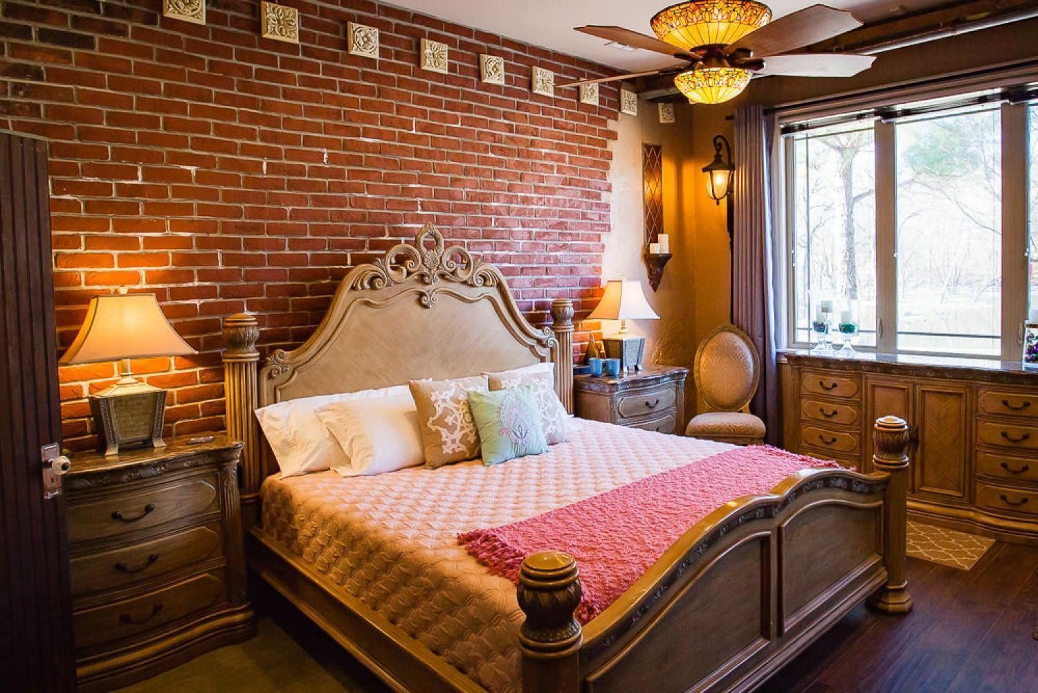 Cozy bedroom with ornate wooden bed, brick accent wall, warm lamp lighting, and large window.