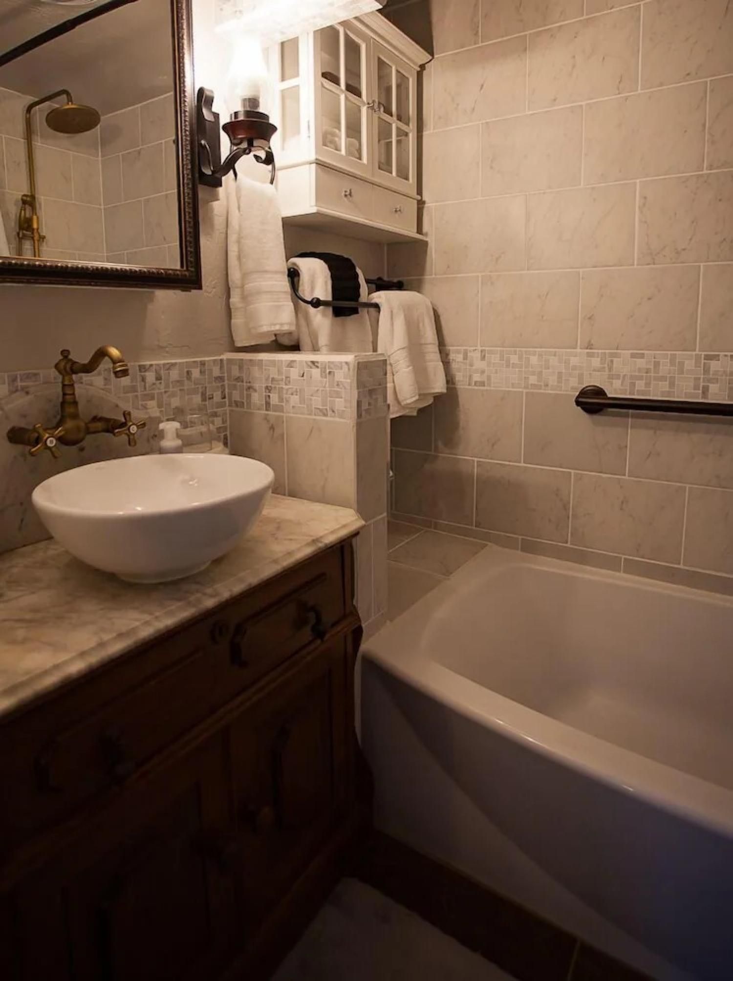 Compact bathroom with stone sink, wood vanity, bathtub, tiled walls, and wall sconce lighting.
