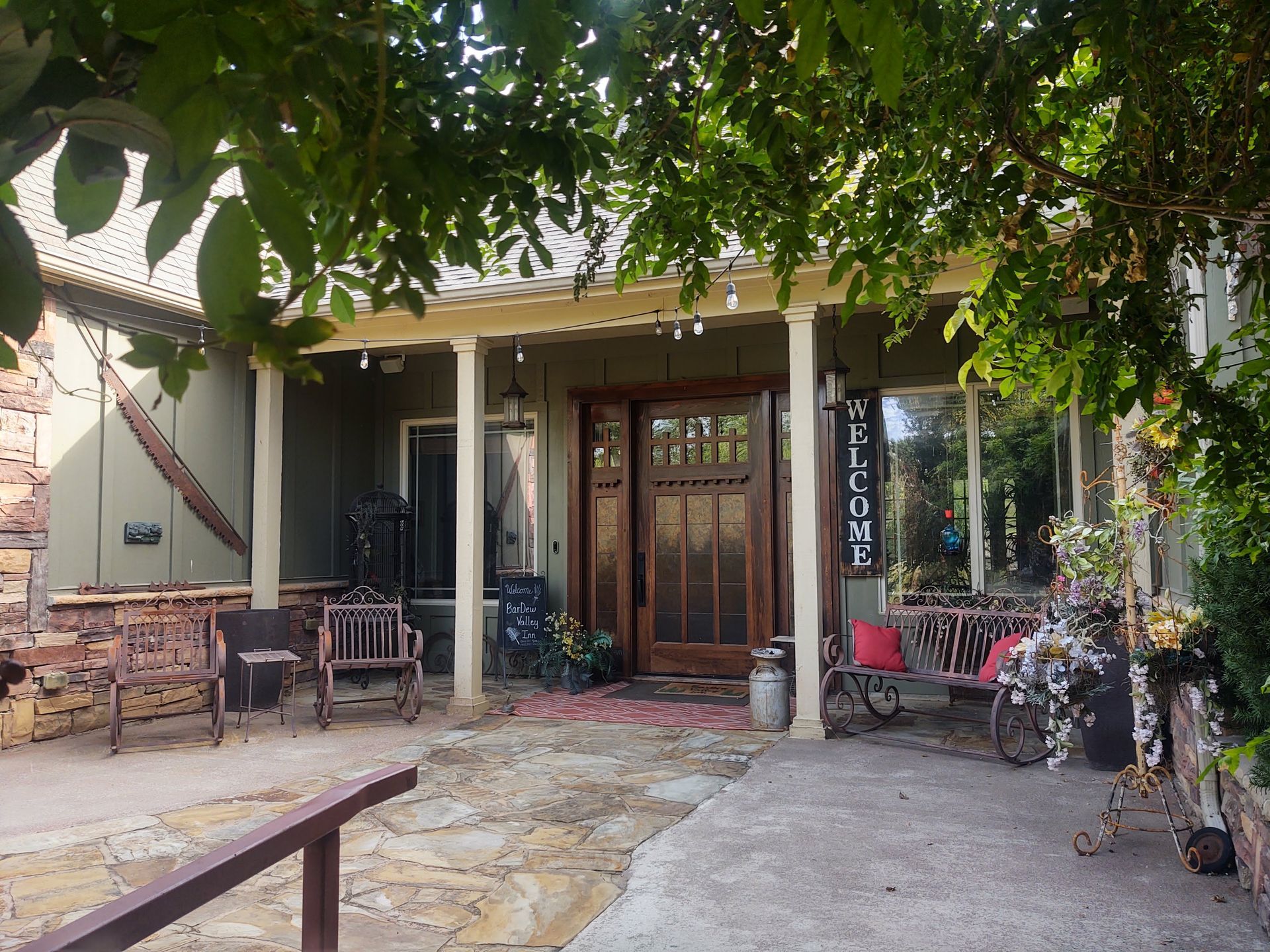 Shaded front porch with chairs, potted plants, and a glass front door under leafy trees