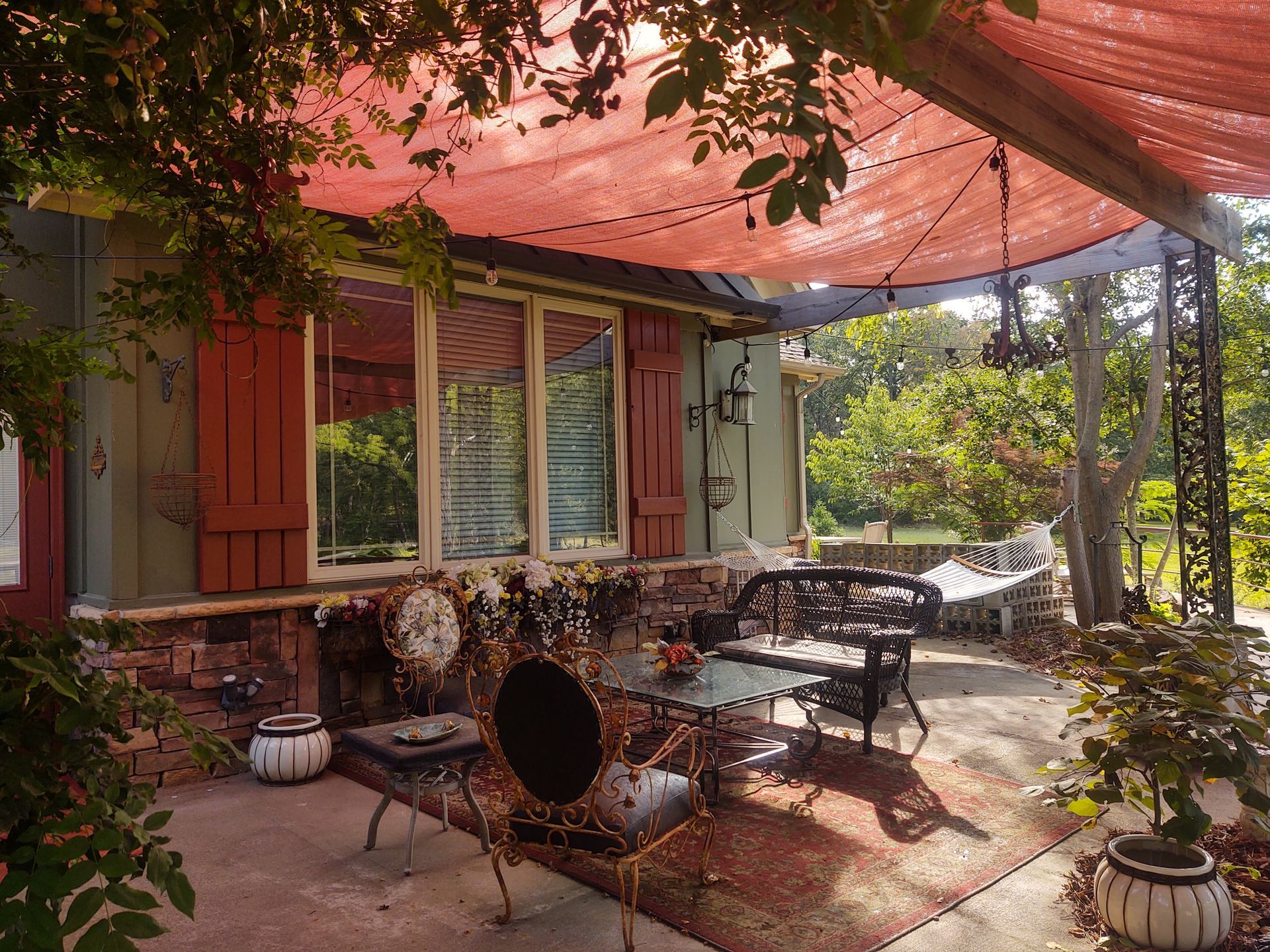 Patio with wicker chairs and table beneath a red canopy beside a house and garden greenery