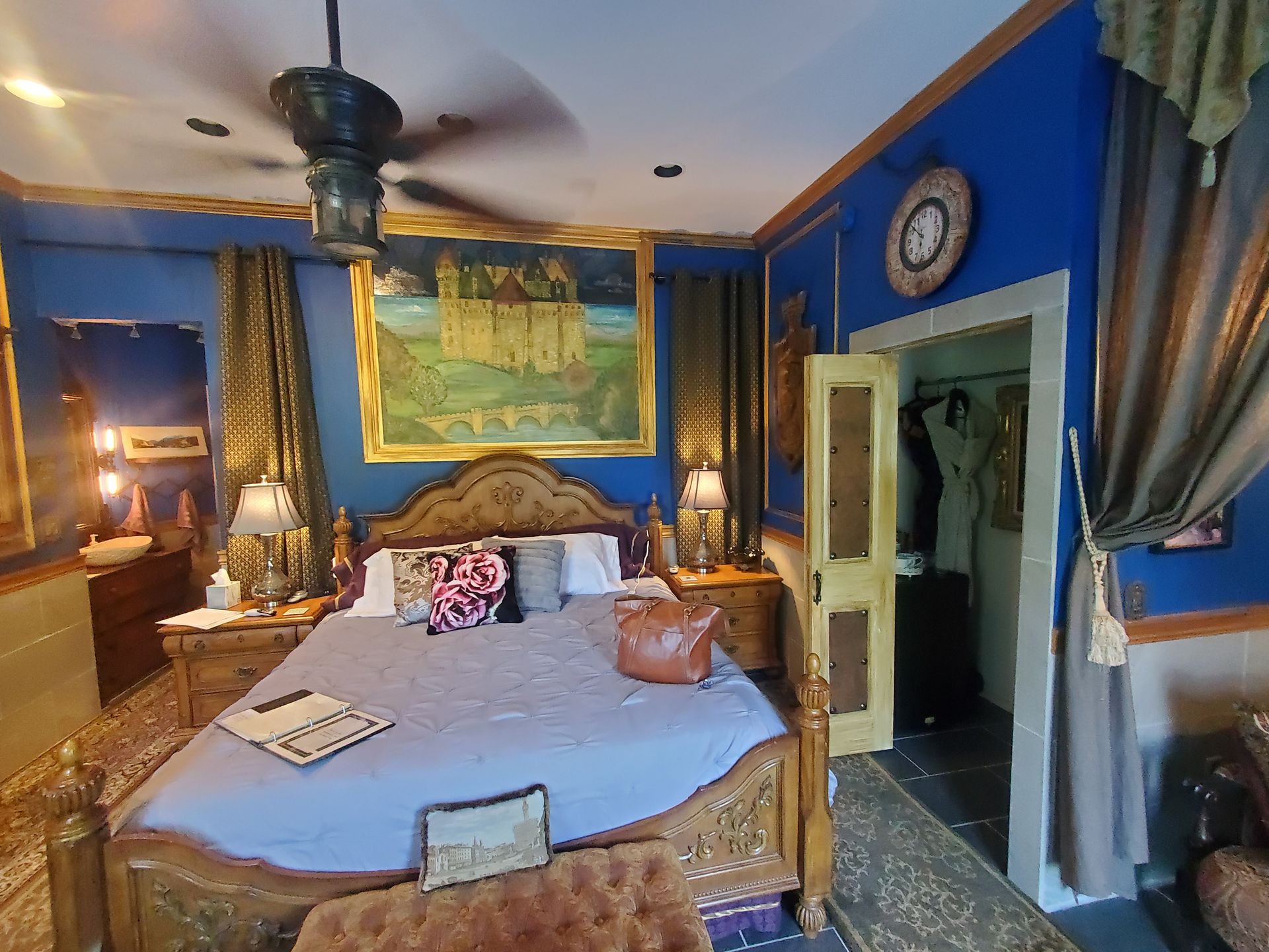 Blue bedroom with ornate carved bed, draped curtains, wall art, mirror, and warm lamp lighting