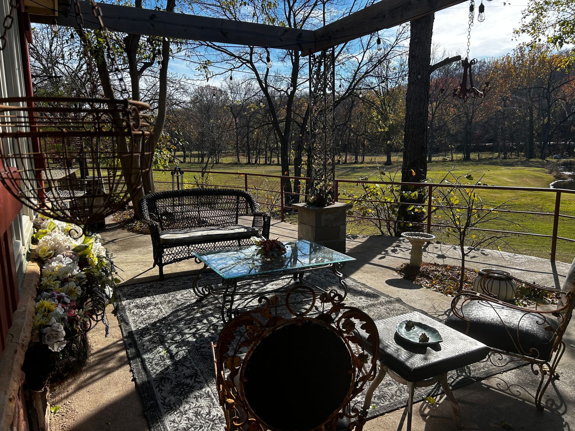 Sunlit patio with wrought-iron furniture, potted flowers, and a view of a grassy yard and trees