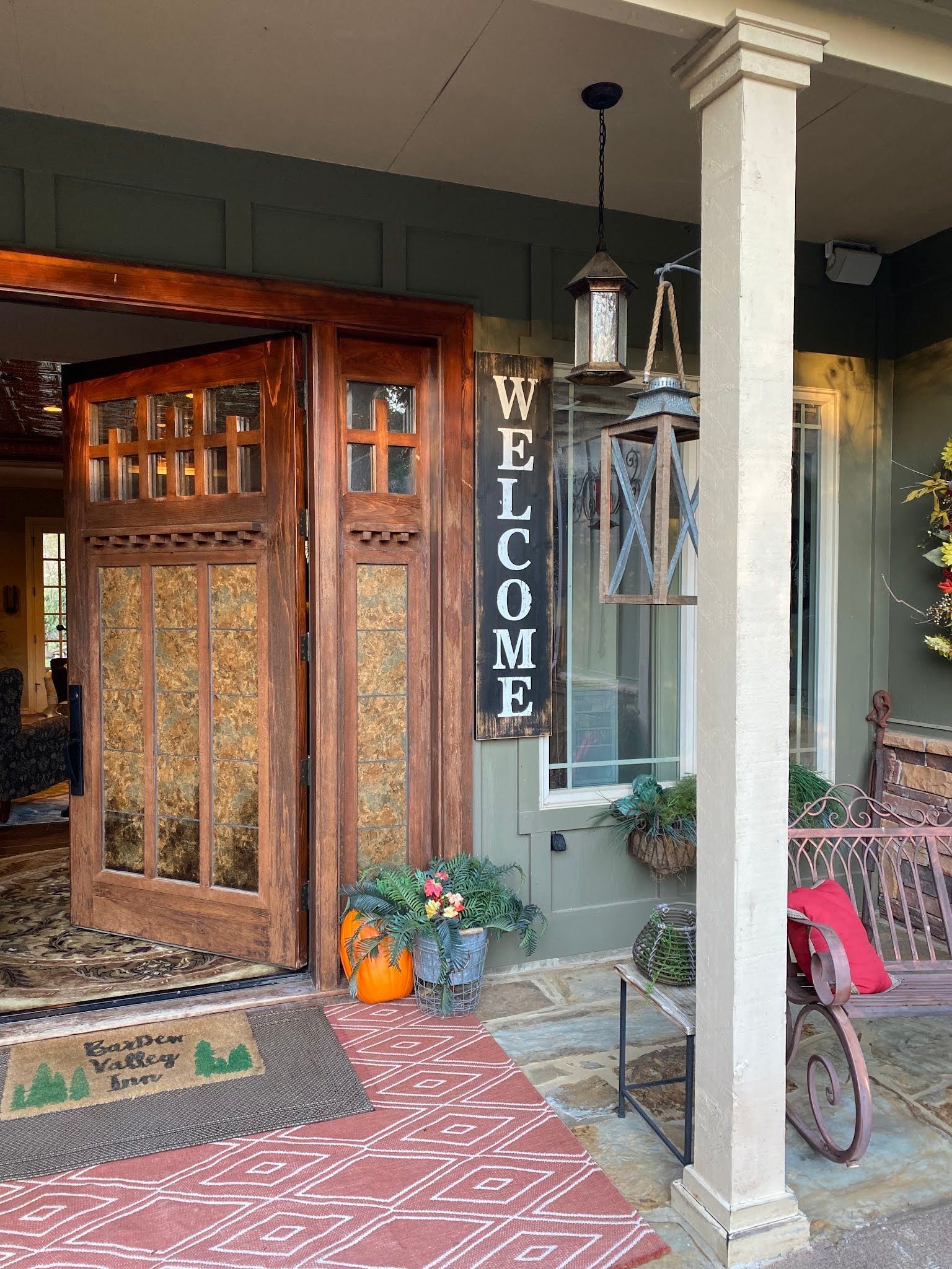 Front porch with open wooden door, welcome sign, potted pumpkins, and a red bicycle beside a column.