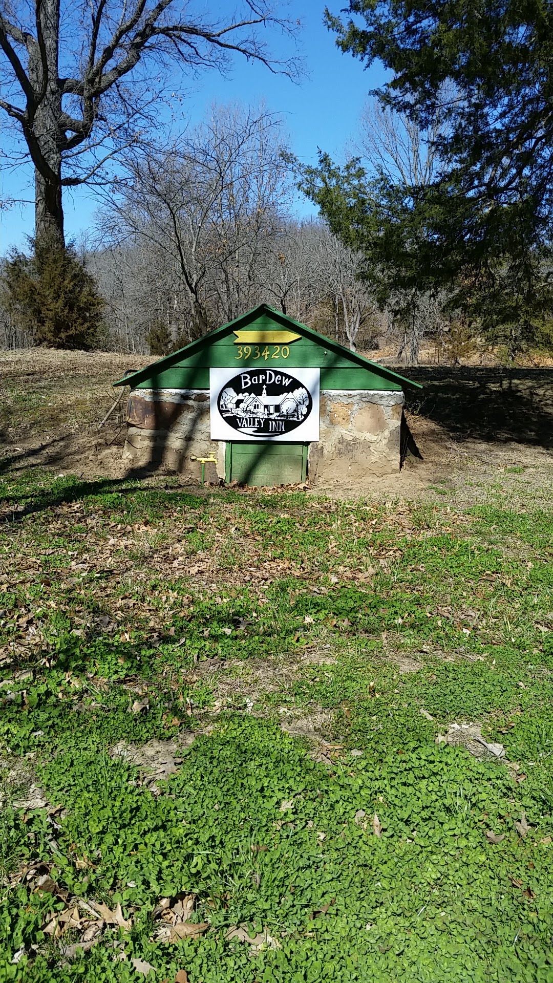 Small green-roofed wooden shelter with a white sign in a grassy, wooded area