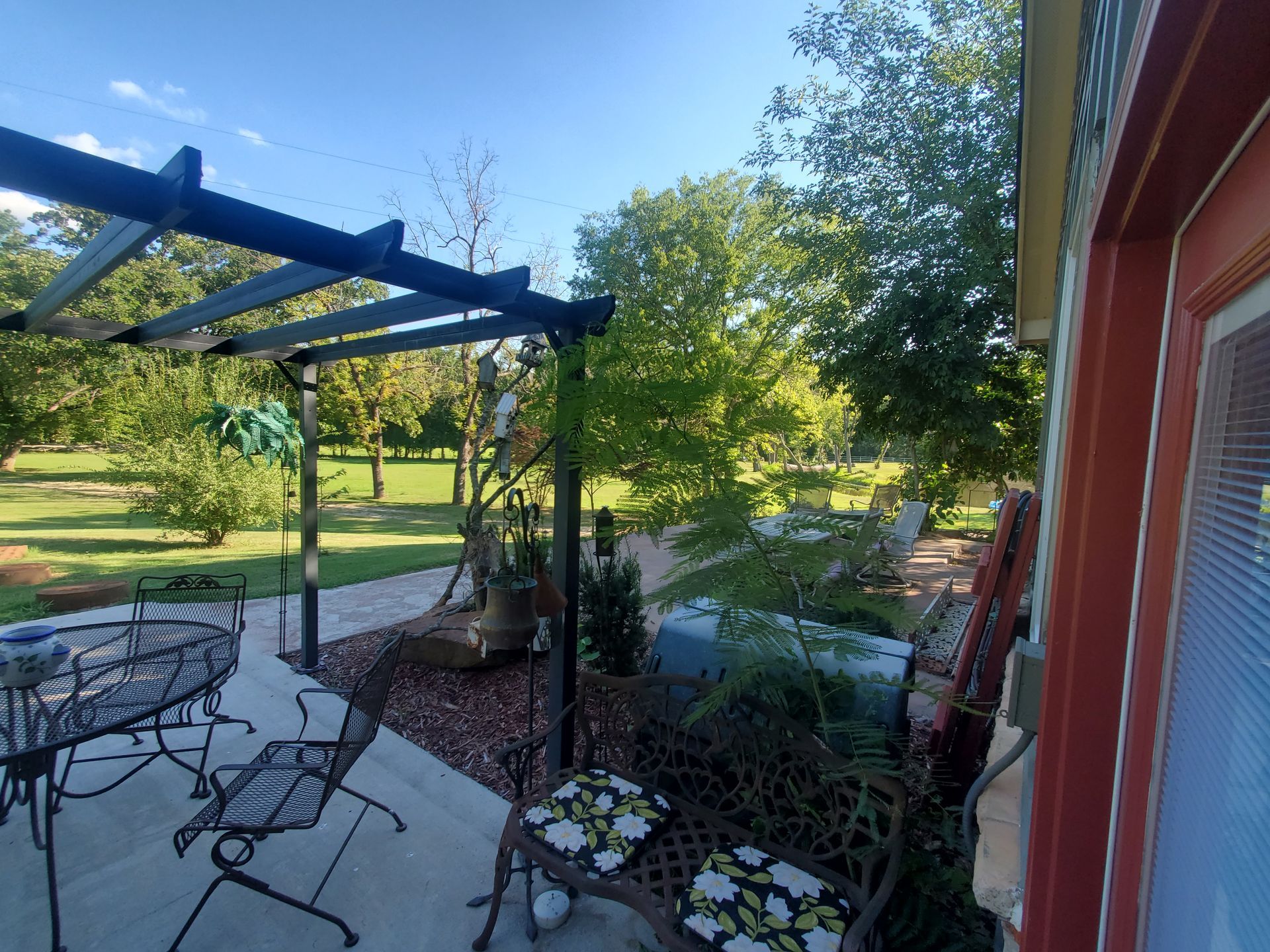 Patio with blue pergola, metal table and chairs, potted flowers, and a sunny backyard view
