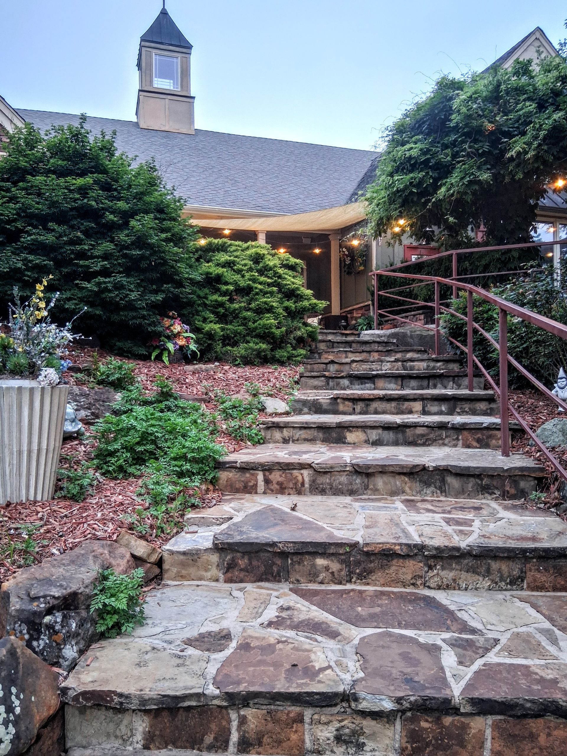 Stone steps leading to a rustic building with a shingled roof, lamppost, and lush greenery
