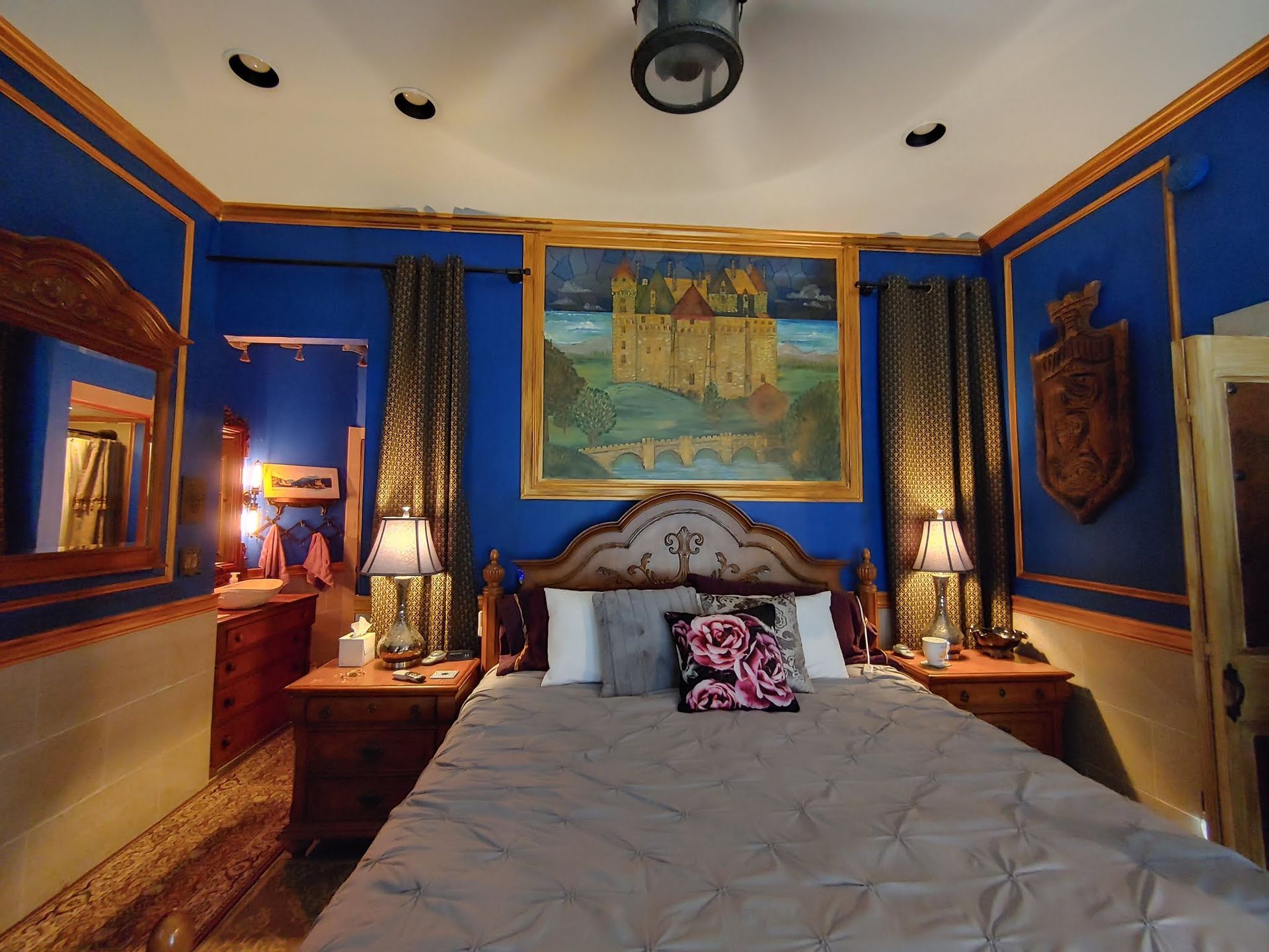 Blue and gold bedroom with a bed, bedside lamps, framed art, and a mirrored wall.