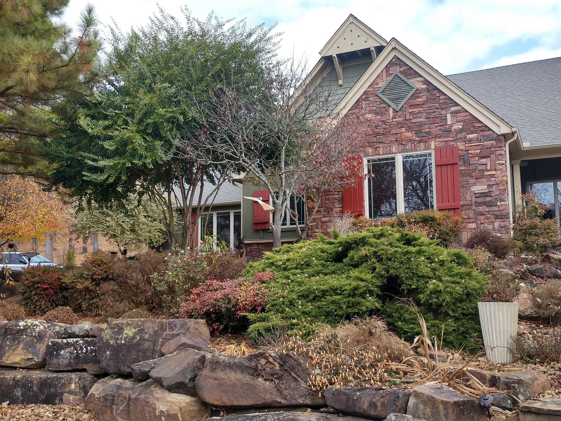 Stone house with red shutters and a landscaped front yard in autumn.