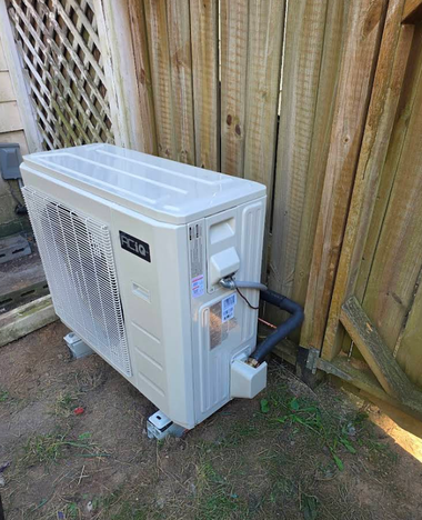 Outdoor wall-mounted air conditioner unit beside a wooden fence on a concrete pad.