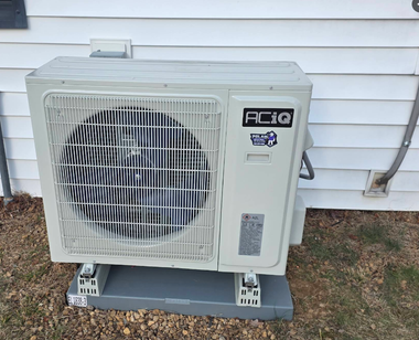 Outdoor AC condenser unit beside a house, white with a large fan grille, on a concrete pad.
