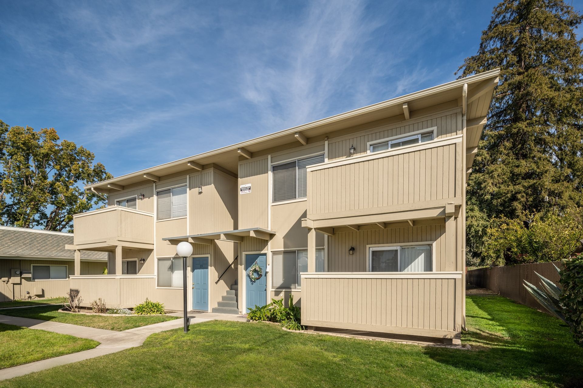 Two-story tan apartment building with blue doors, balconies, and green lawn under a blue sky.
