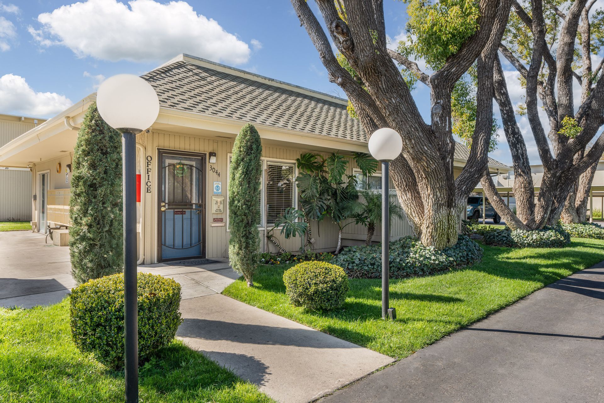 Small, beige building with blue door, white globe lights, and green landscaping under a sunny sky.