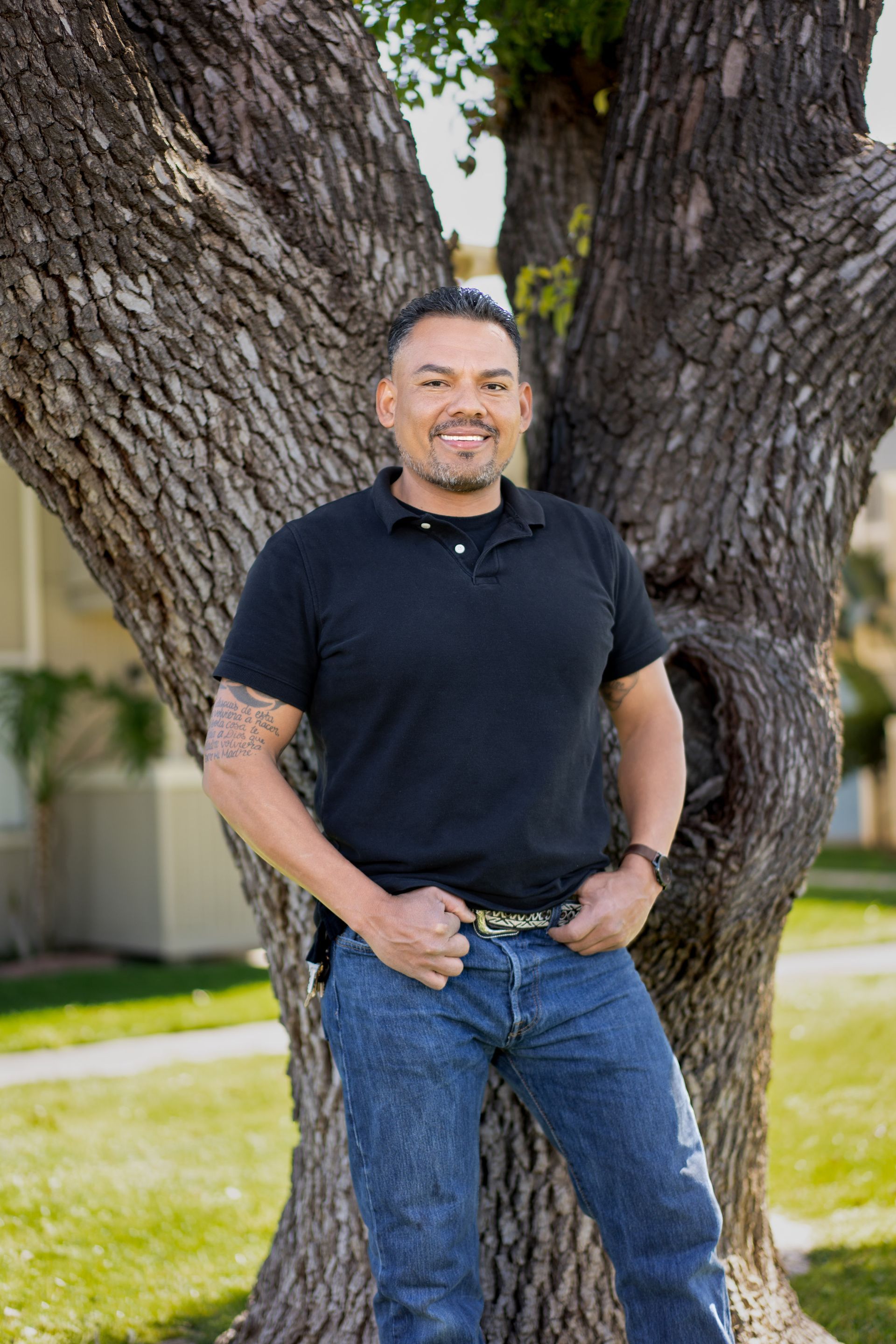 Man in black shirt and jeans leans against a tree, smiling in a sunny outdoor setting.
