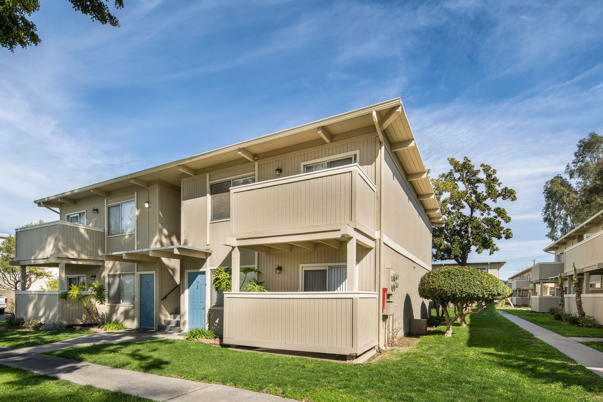 Two-story tan apartment building with small balconies and green grass, under a blue sky.