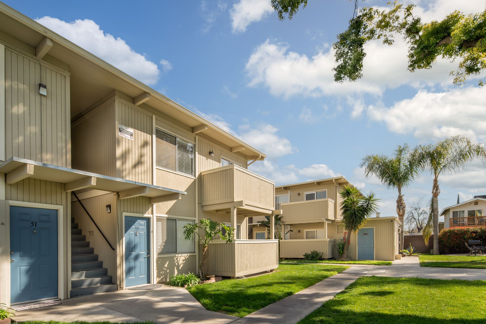 Apartment buildings with beige siding, blue doors, and green lawns under a blue sky with some trees.