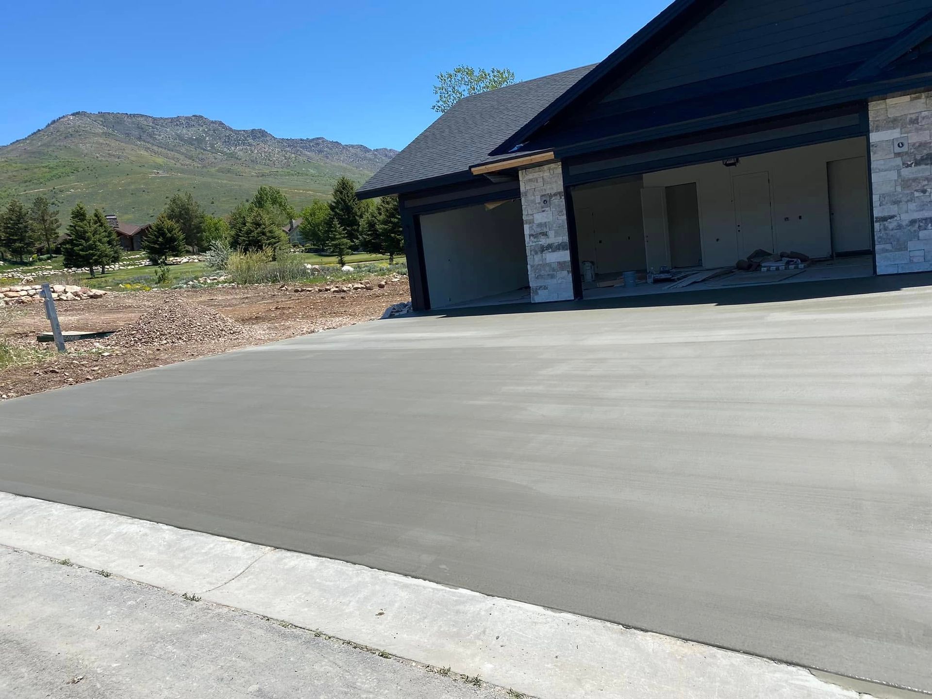 A new, smooth concrete driveway leads to a modern garage under a mountain backdrop on a sunny day.