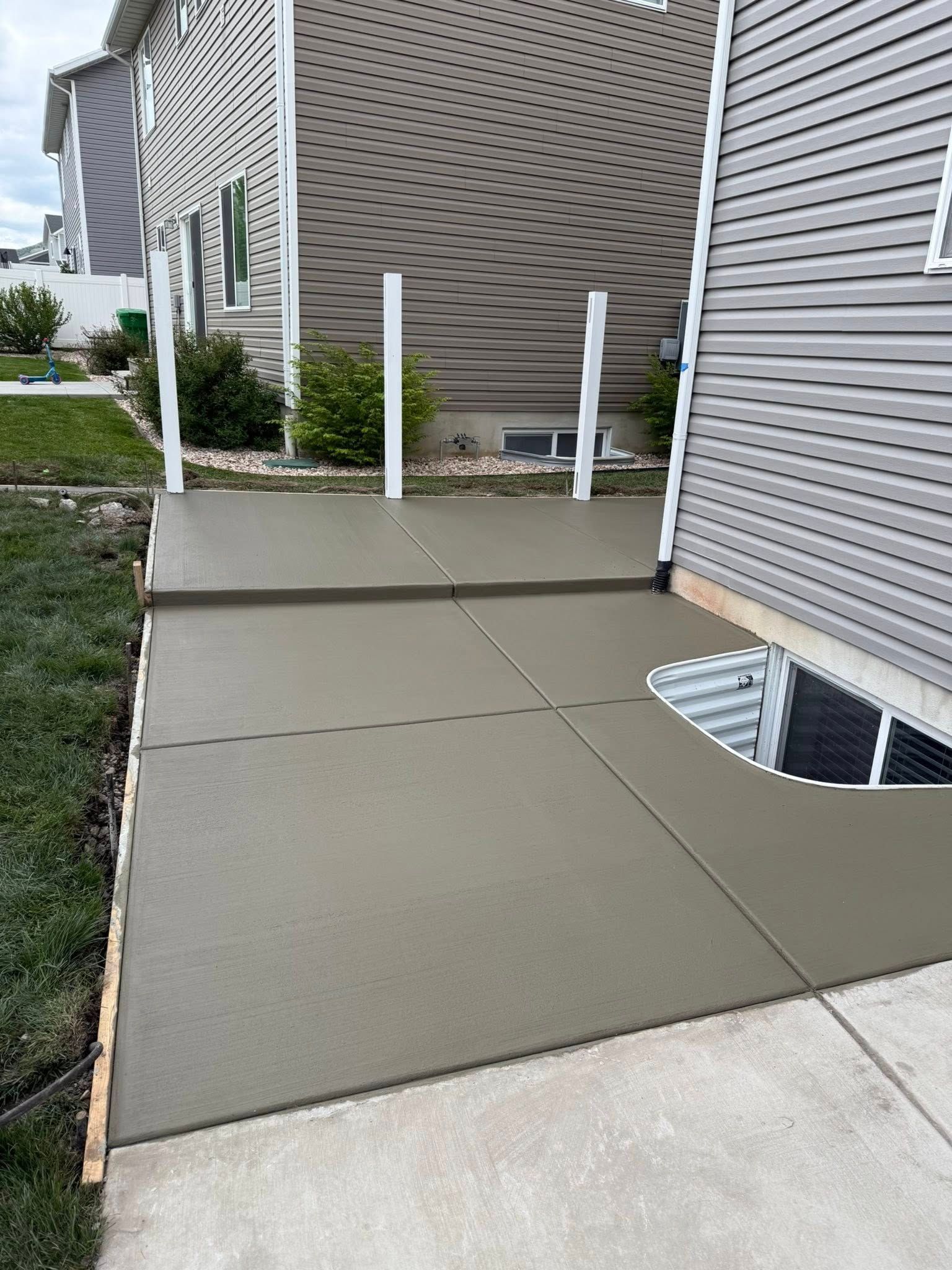 Freshly poured gray concrete patio extension adjacent to a house with siding and a basement window well.