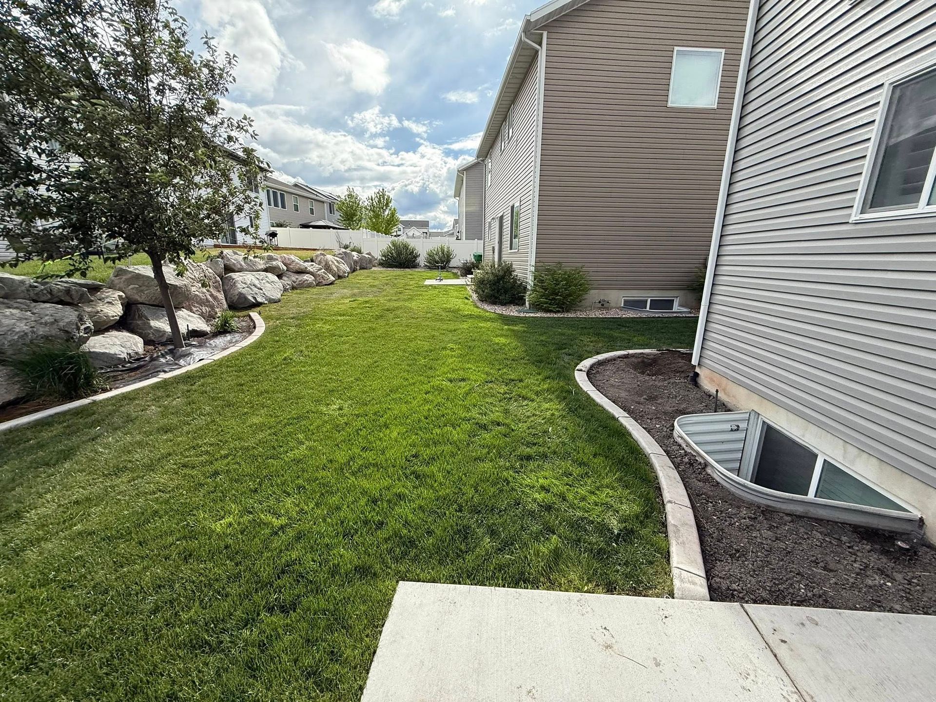 A side yard with a manicured lawn, a large rock retaining wall, and a window well along the side of a house.