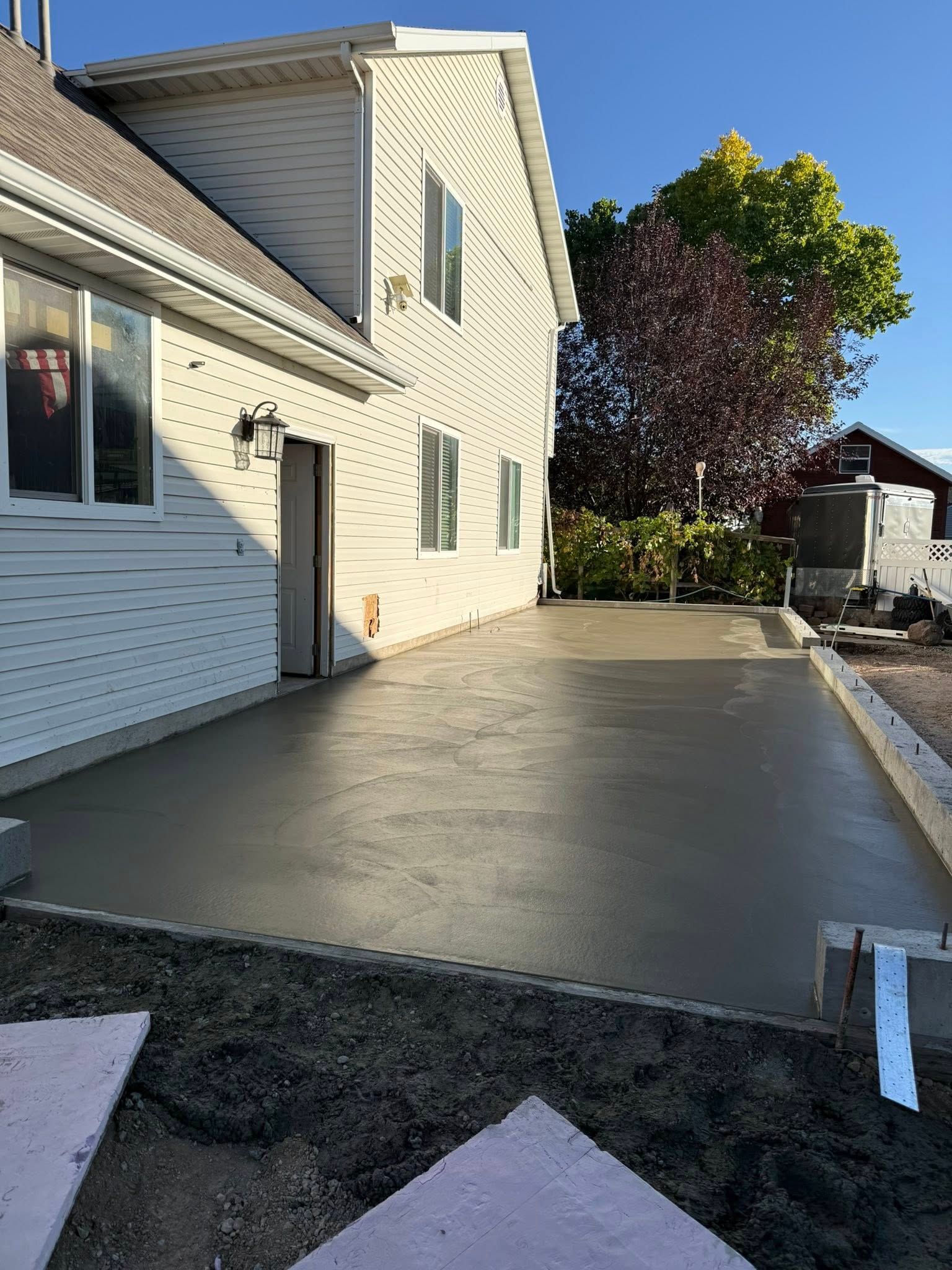 A freshly poured concrete patio adjacent to a light-colored two-story house under a clear blue sky.