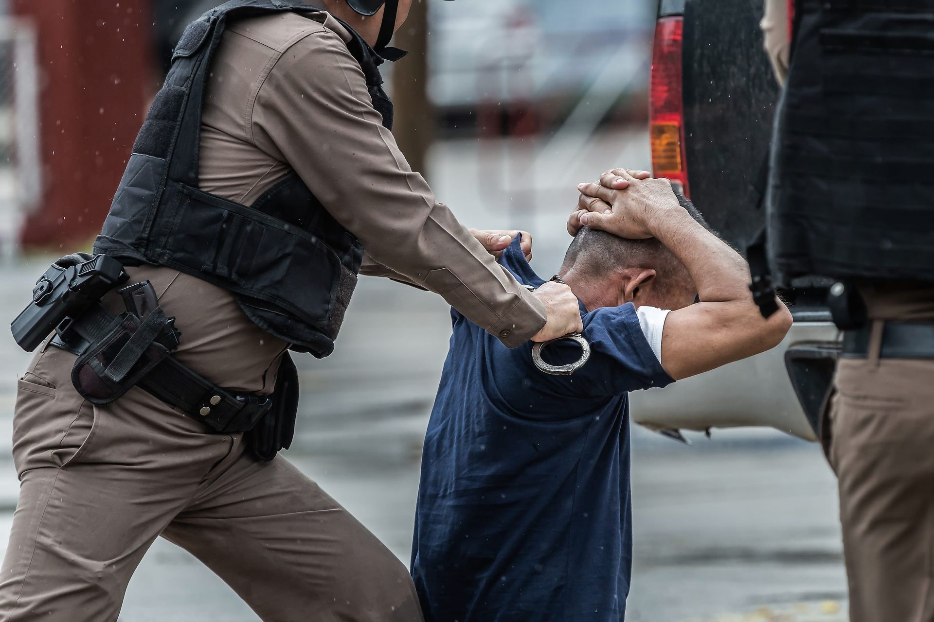 A law enforcement officer in tan uniform and tactical vest detains an individual by applying handcuffs.