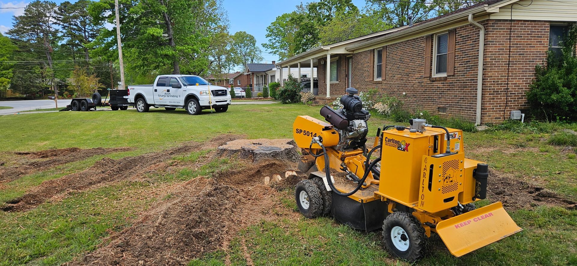 Yellow stump grinder in front yard near a brick house. A white pickup truck is parked in the background.