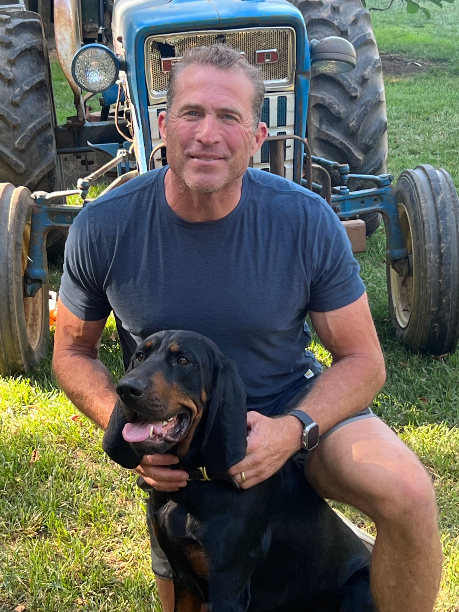 A man is kneeling down with a dog in front of a tractor.